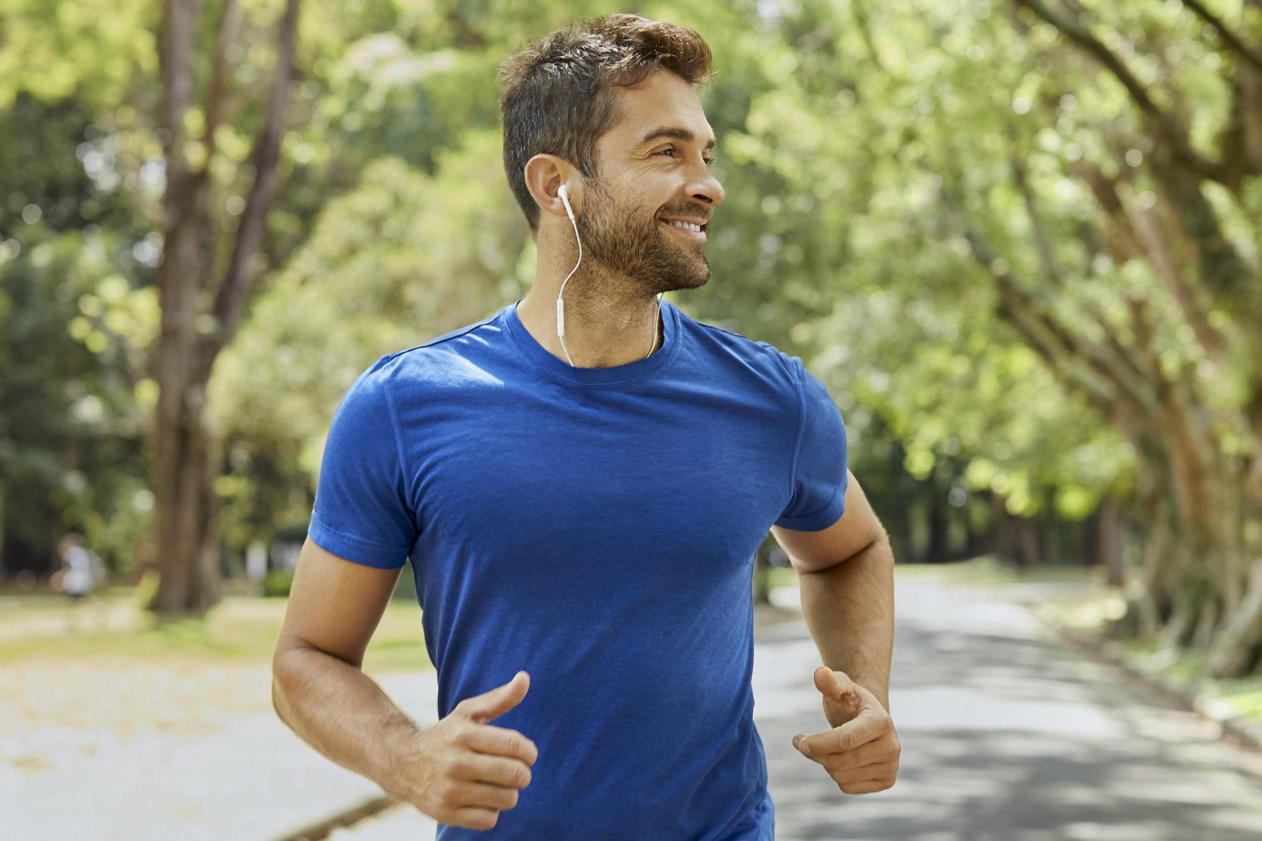 مردی خندان با تی شرت آبی در یک پارک در حال دویدن است.