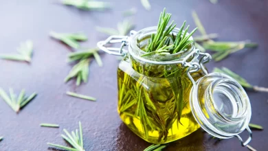 یک شیشه روغن رزماری در میان برگ های سبز این گیاه روی یک میز بنفش