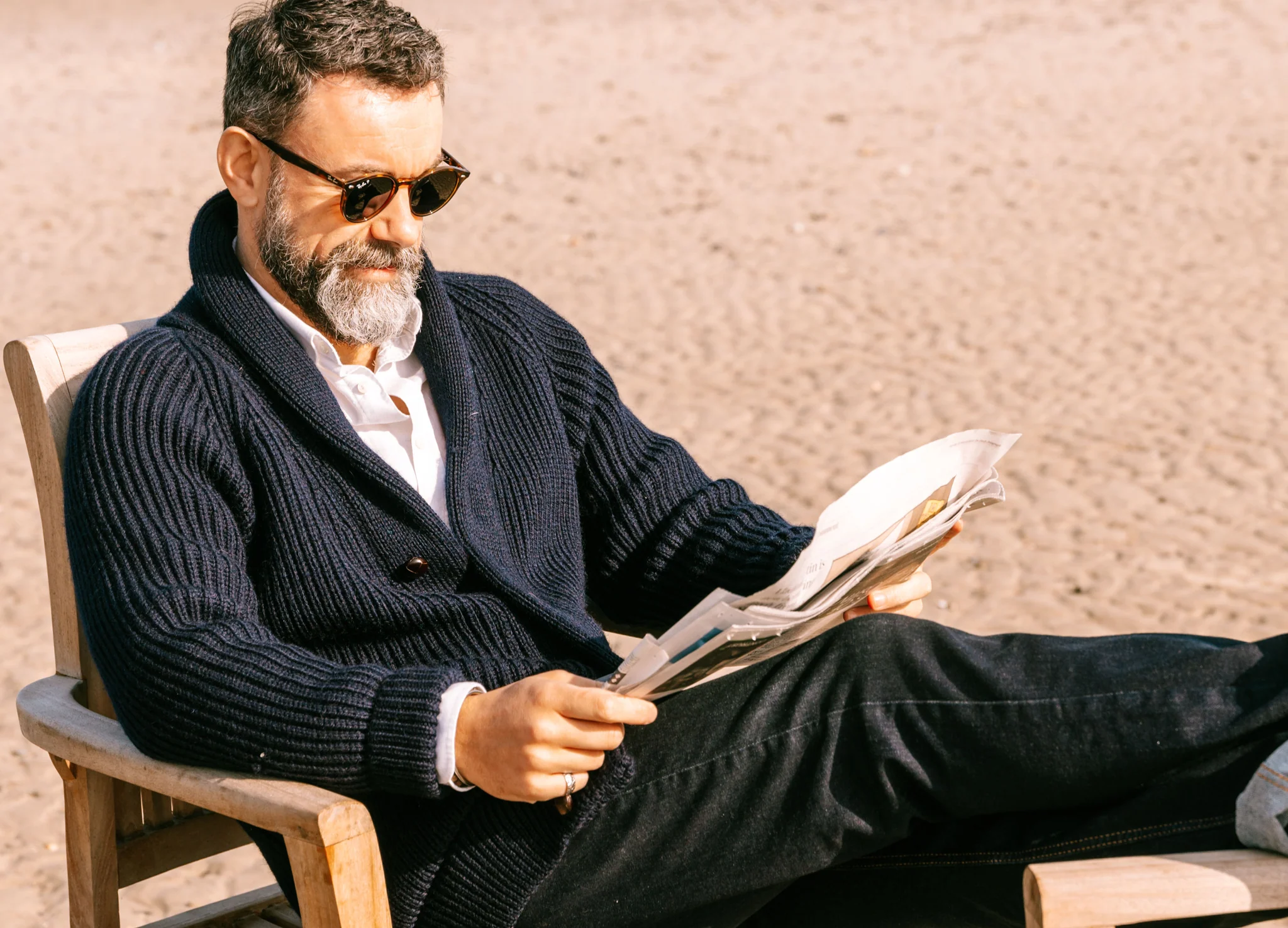 یک مرد جوان که عینک آفتابی به چشم دارد و ژاکت یقه شال پوشیده روی صندلی در حال خواندن روزنامه است.