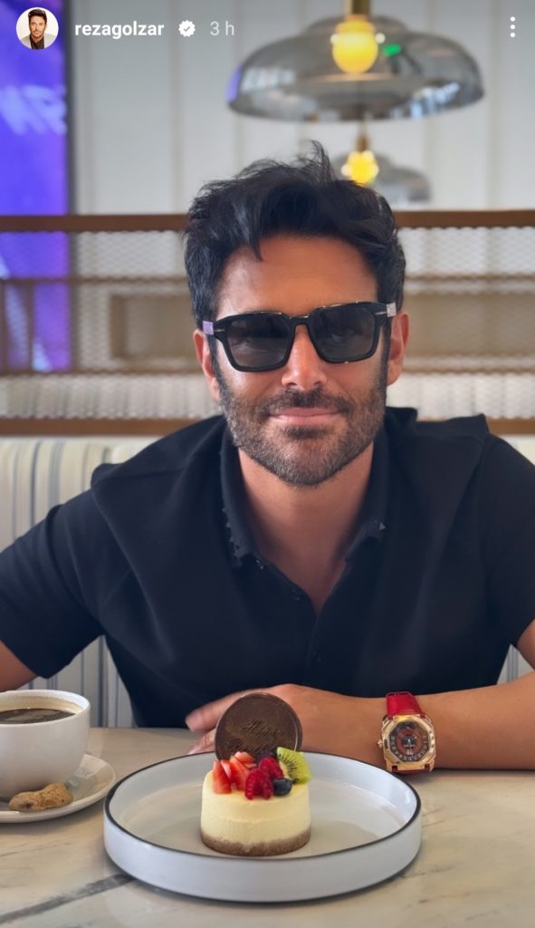 محمدرضا گلزار با عینک آفتابی، تیشرت سرمه ای و ساعت قرمز پشت یک کیک کوچک